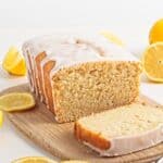 Lemon loaf cake with icing is sliced on a wooden board, surrounded by whole and sliced lemons.