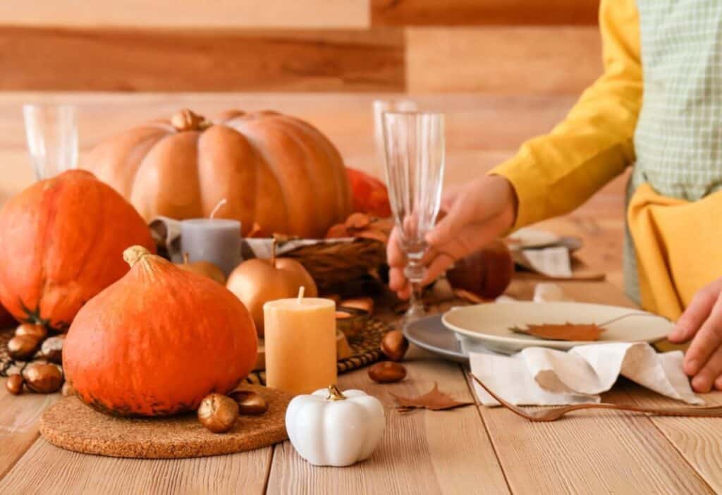 A table decorated for autumn with pumpkins, candles, and leaves. Person setting a table with glassware and plates.