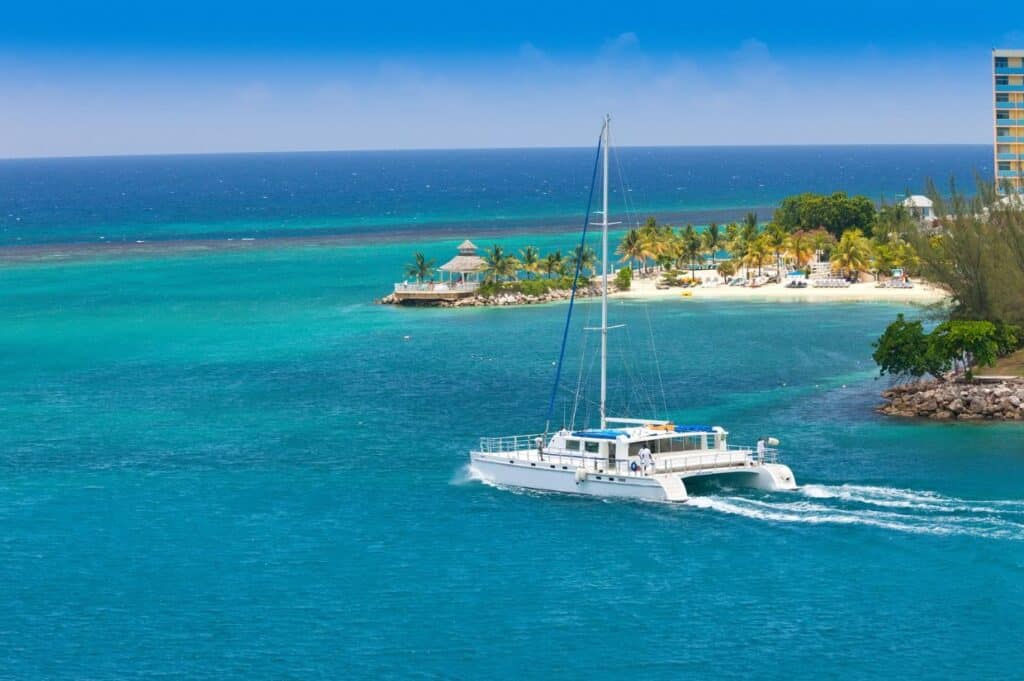 A white catamaran sails on clear turquoise waters, embodying the essence of sailing the Caribbean near a tropical island with palm trees and a building in the background.