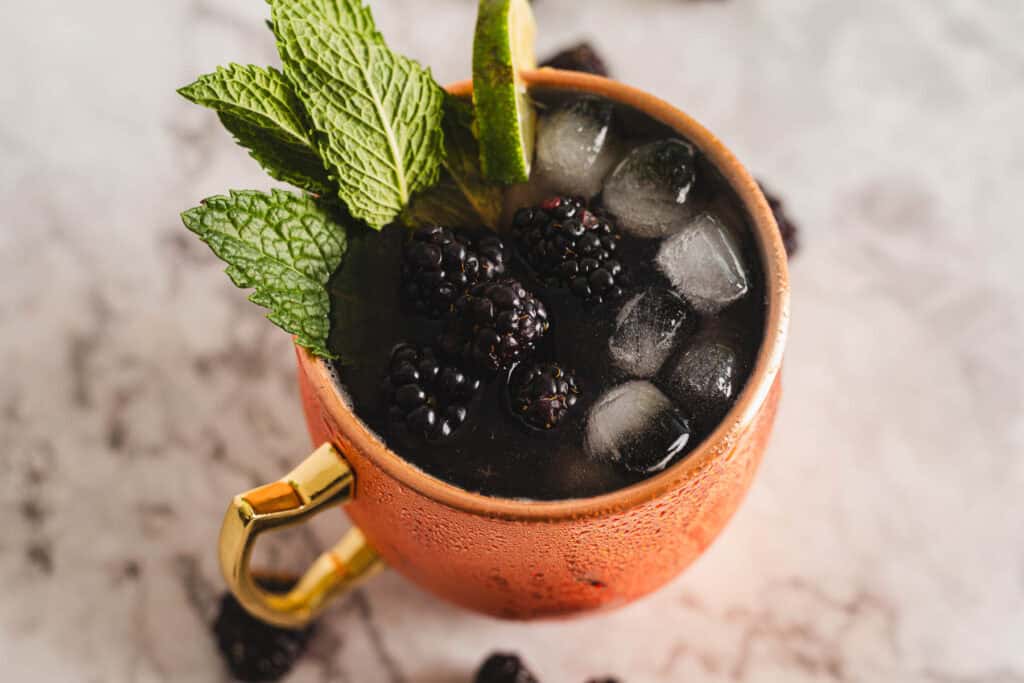 A copper mug filled with a dark beverage, ice cubes, blackberries, and garnished with mint leaves and a lime wedge, placed on a marble surface.