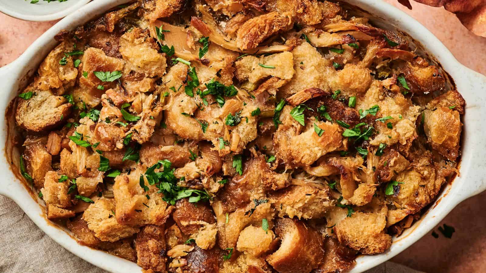 A baked dish of golden-brown stuffing with pieces of bread, herbs, and a crispy top garnished with chopped parsley in a white oval dish.