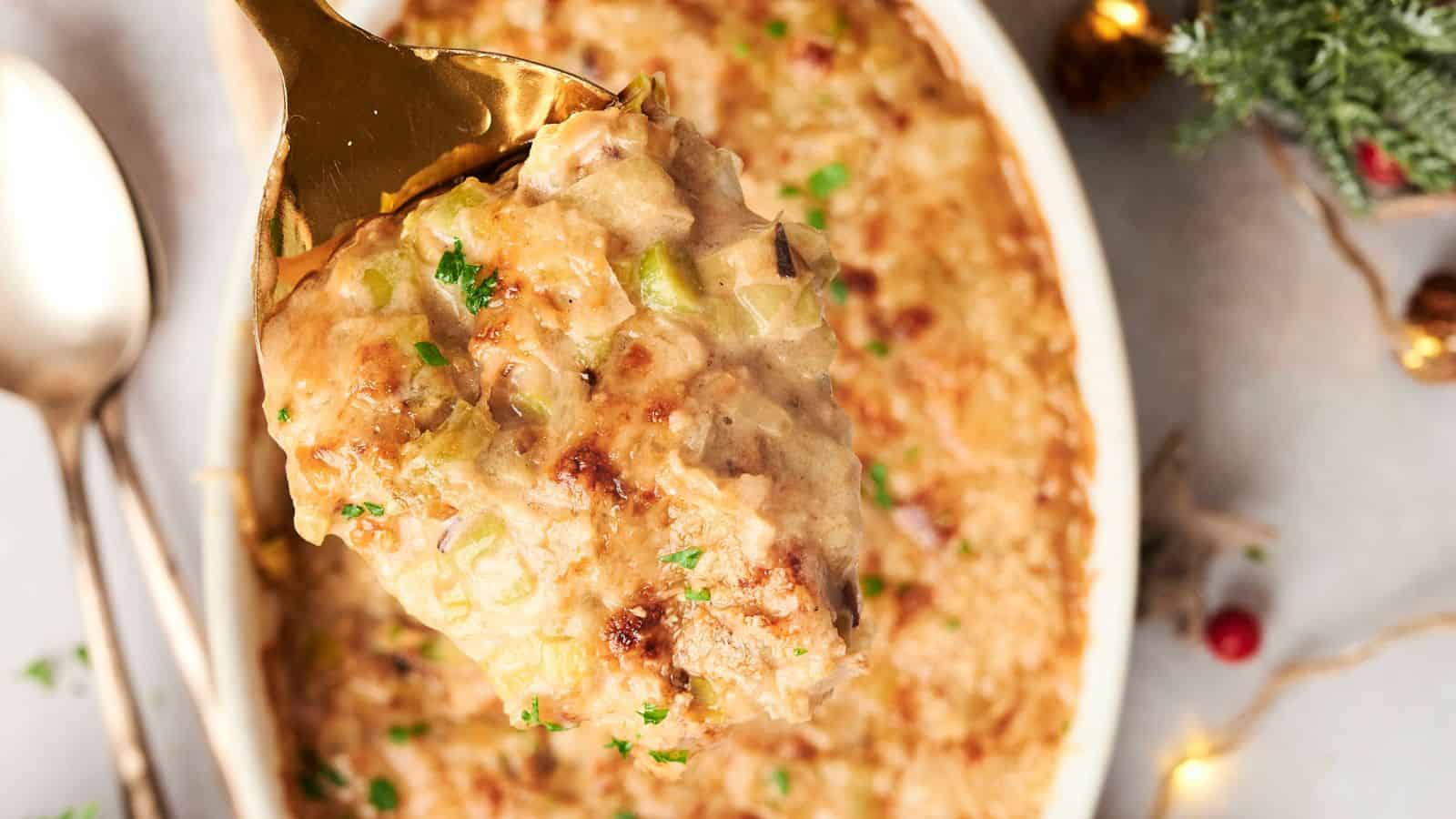 A close-up of a spoon scooping a portion of creamy baked casserole topped with herbs, with a dish of the casserole and a spoon in the background.