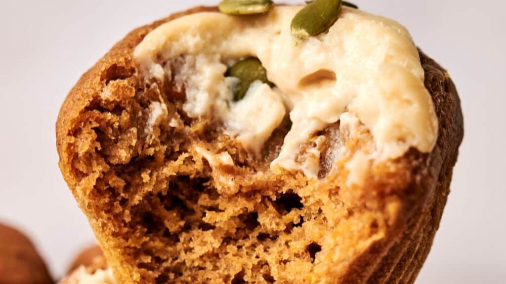 Close-up of a bitten muffin topped with cream and pumpkin seeds.