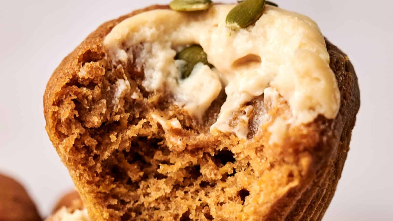 Close-up of a muffin with a bite taken out, topped with cream and pumpkin seeds.