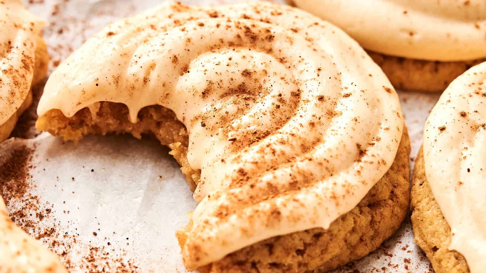 Close-up of a frosted cookie with a bite taken out, sprinkled with cinnamon, on a parchment surface.