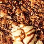 Close-up of a baked dessert topped with chopped pecans, ice cream, and caramel drizzle in a white dish.