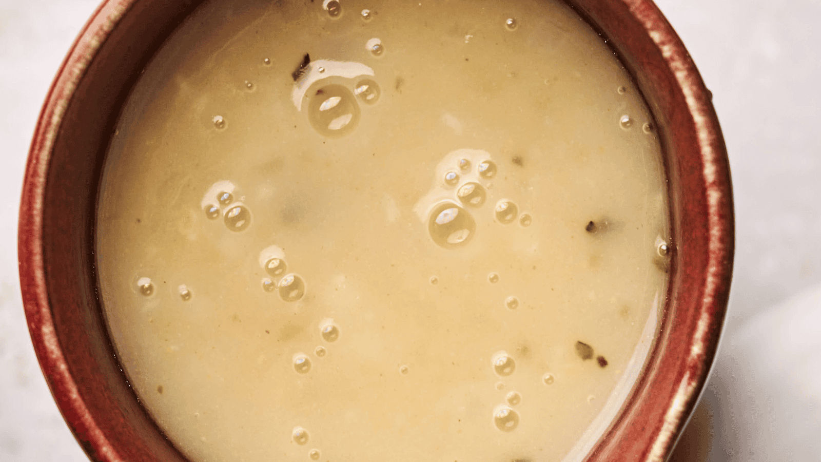 Close-up of a bowl filled with creamy turkey gravy soup, featuring small bubbles and herbs on the surface. The bowl has a reddish-brown rim.