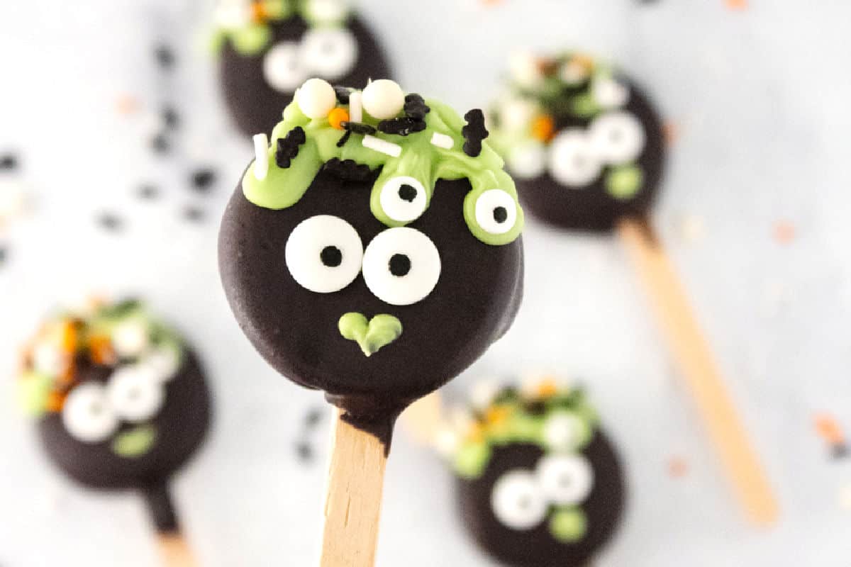 Chocolate cookie pop decorated with green icing, googly eyes, and sprinkles, resembling a monster.