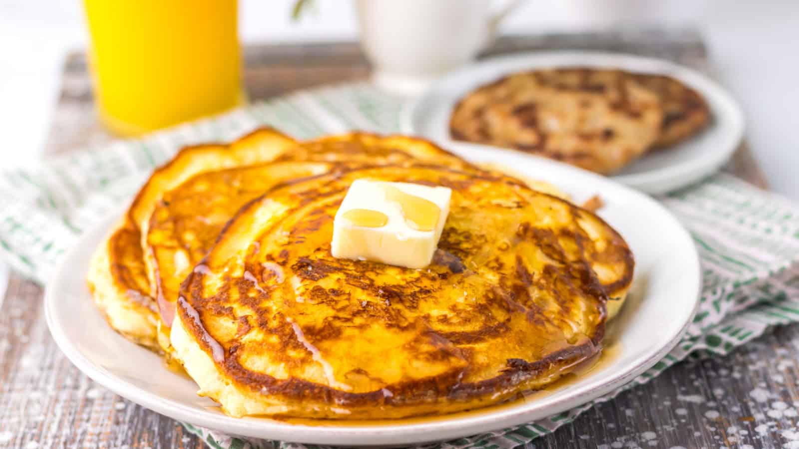 Plate of pancakes topped with butter and syrup, with a drink and a cookie on a table covered by a patterned cloth.