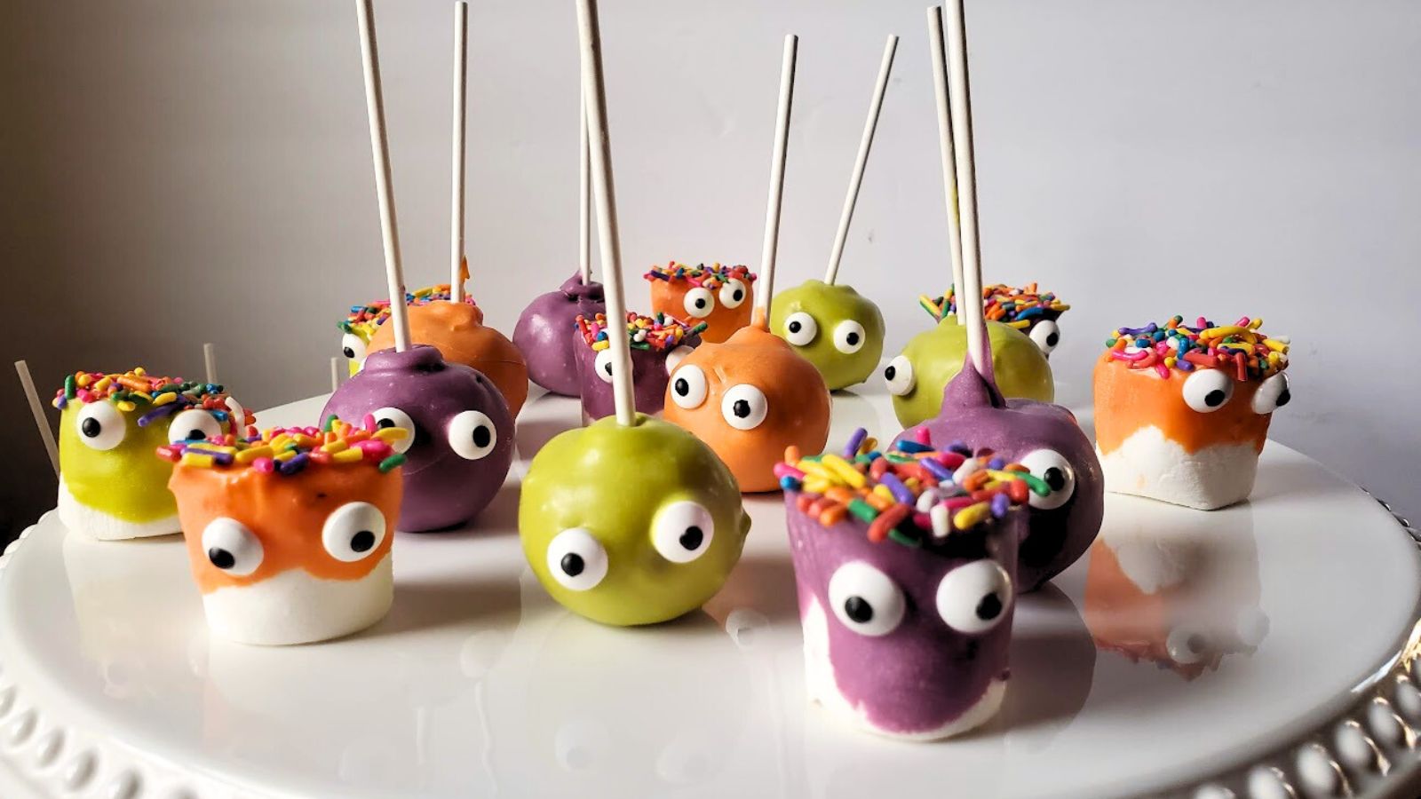 Colorful cake pops and marshmallows with googly eyes and sprinkles on a white plate.