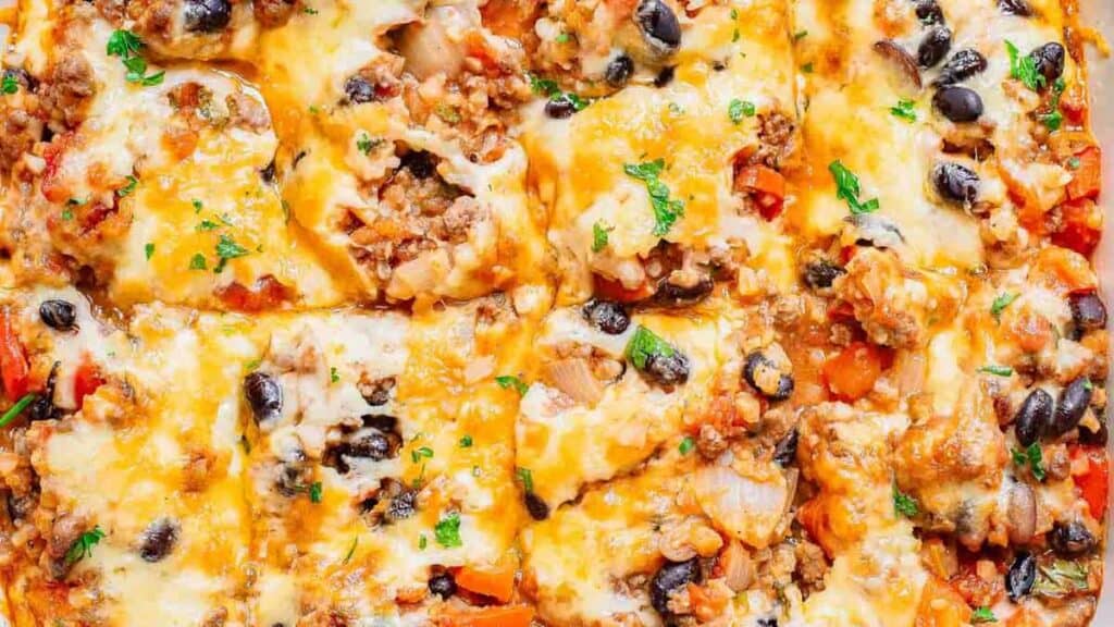 Close-up of a cheesy taco casserole with layers of melted cheese, seasoned ground beef, black beans, peppers, and onions, garnished with chopped parsley.