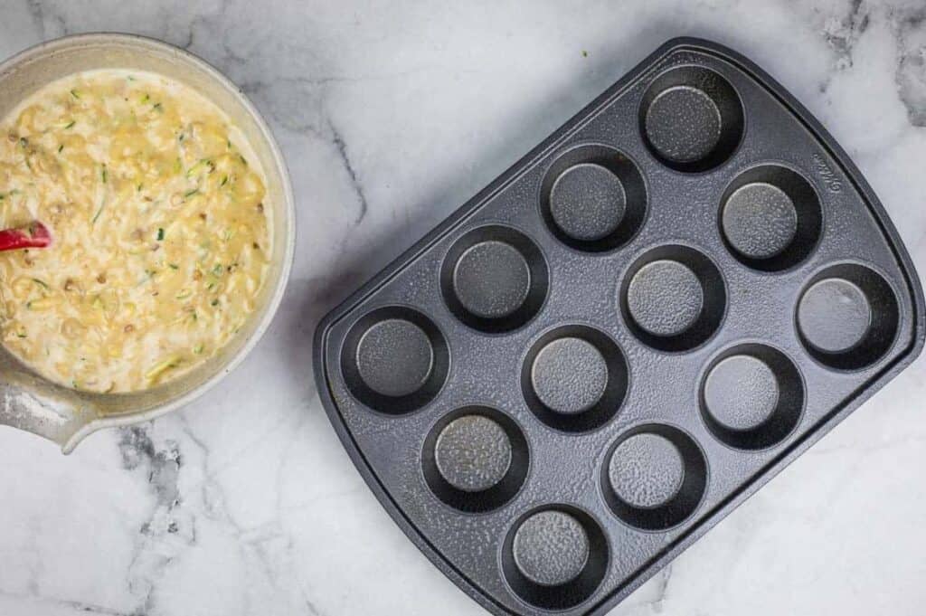 Empty muffin tray next to a bowl of batter on a marble countertop.