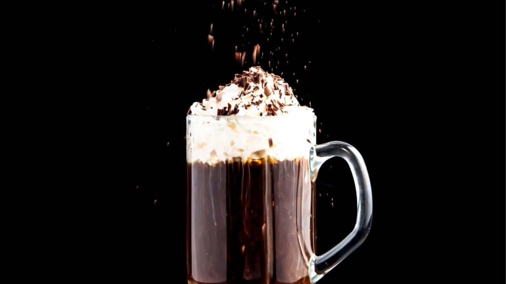 A glass mug filled with frothy coffee topped with whipped cream and chocolate shavings against a black background.