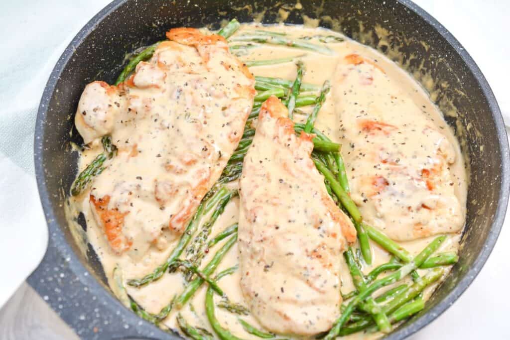 Chicken breasts in a creamy sauce with herbs and asparagus in a frying pan.