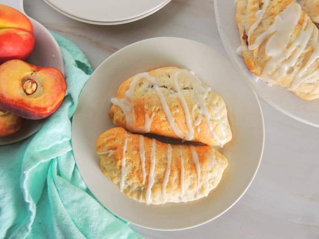 Two iced turnovers on a white plate, next to a halved peach on a light green cloth.