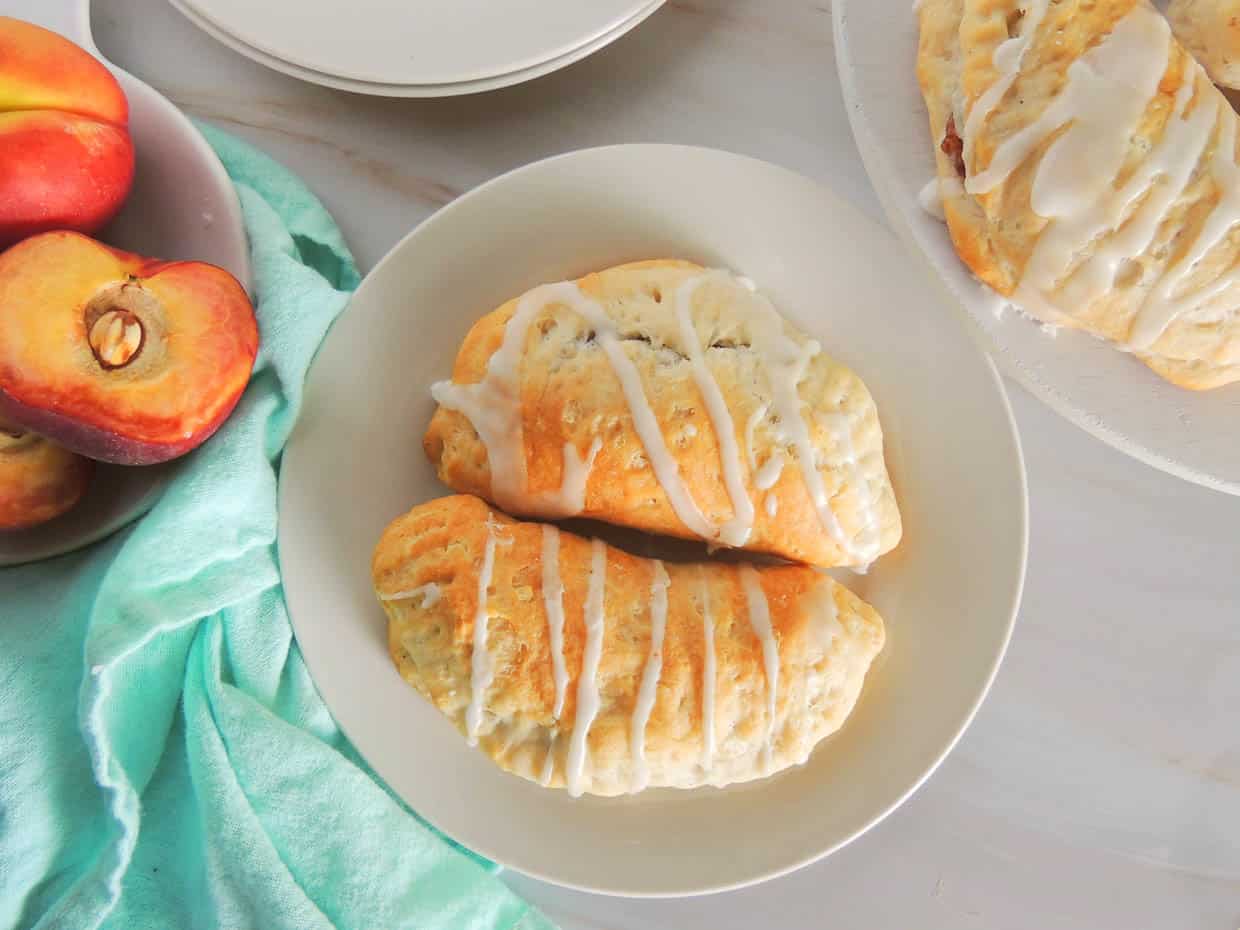 Two glazed peach hand pies on a plate next to fresh peaches and a teal napkin.