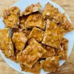 A white plate filled with pieces of peanut brittle on a wooden table.