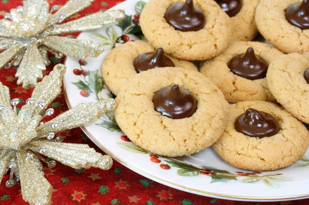 A plate of peanut butter blossom cookies with chocolate centers, perfect for a holiday cookie exchange, rests on a festive red and gold tablecloth adorned with decorative silver snowflakes.