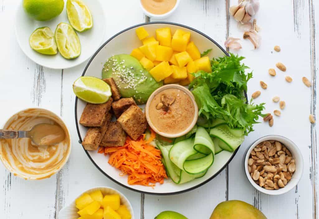 A salad bowl with tofu, mango, lettuce, cucumber, carrots, avocado, and lime, served with peanut sauce. Surrounded by lime, mango, peanuts, garlic, and more sauce on a white background.