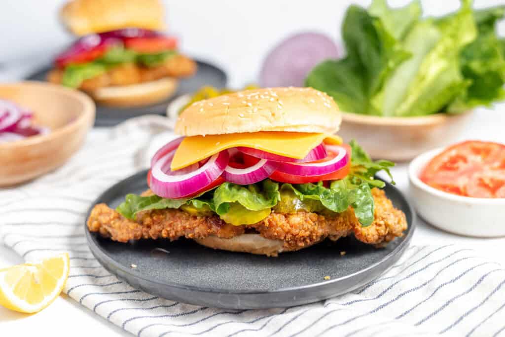 Fried chicken sandwich with cheese, lettuce, and red onion on a black plate. Sliced tomatoes and lettuce in the background.