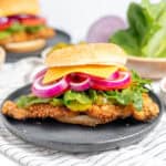 Fried chicken sandwich with cheese, lettuce, and red onion on a black plate. Sliced tomatoes and lettuce in the background.