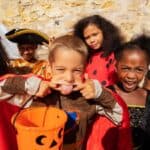 Children in Halloween costumes, including a knight with a pumpkin bucket brimming with treats, a pirate, a ladybug, and a skeleton, stand in front of a stone wall.