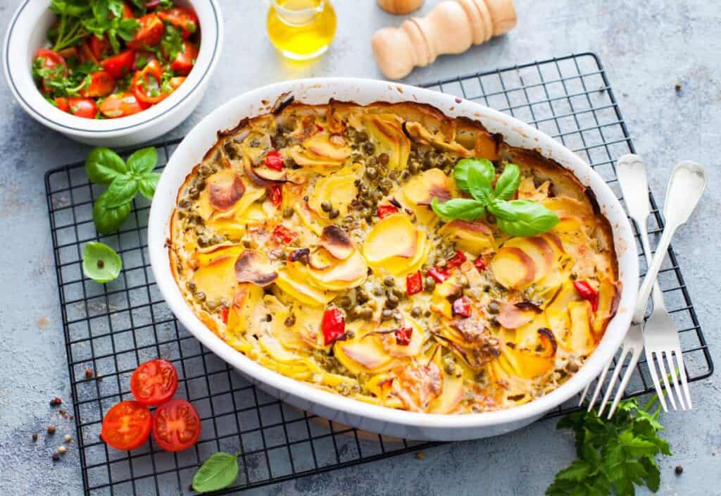 A delightful return of the casserole features a medley of baked vegetables, with sliced potatoes and red peppers layered in a white dish. Garnished with fresh herbs, it's paired perfectly with a bowl of tomato salad and a bottle of oil for an exquisite meal.