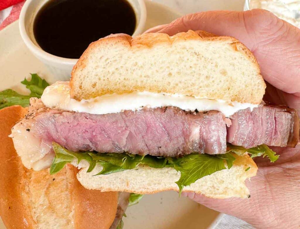 A hand holds a sandwich with sliced steak, lettuce, and a creamy sauce. A small bowl of dark sauce is in the background.