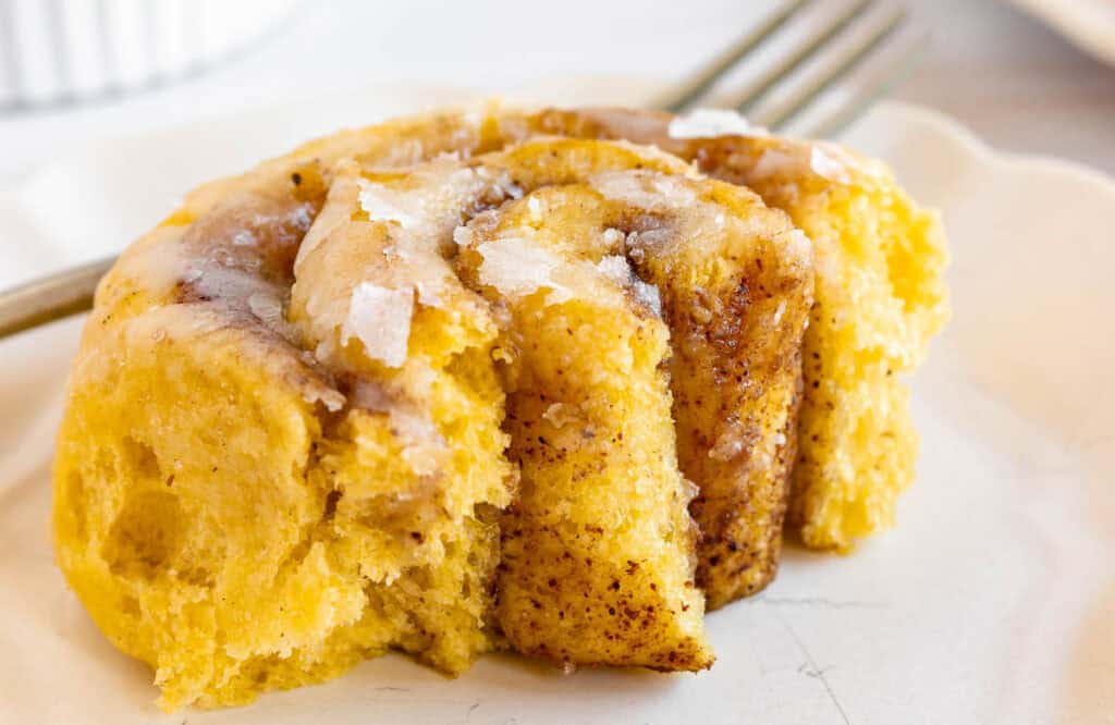 Close-up of a cinnamon roll with icing on a white plate, partially eaten, with a fork nearby.