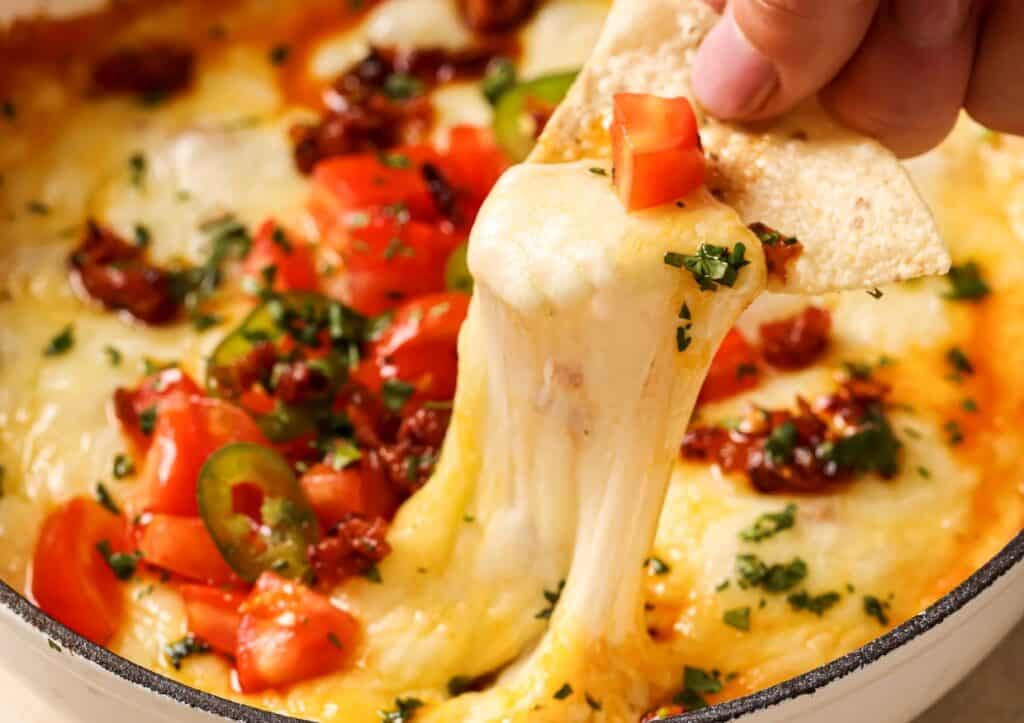 A hand holds a tortilla chip with melted cheese, garnished with diced tomatoes and herbs, being lifted from a cheese dip.