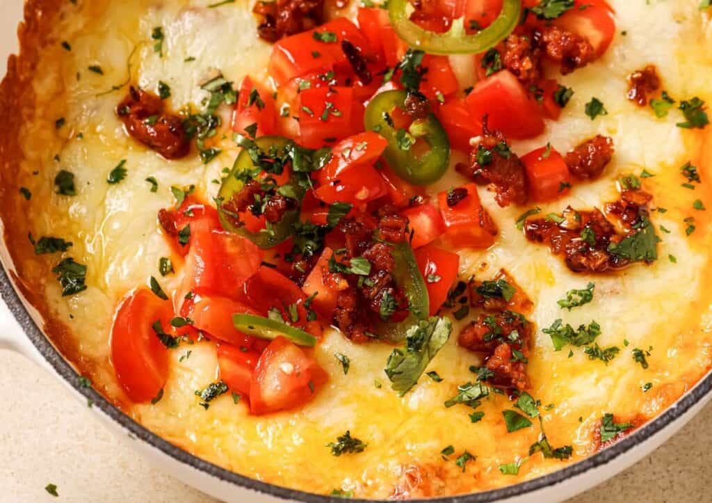 A baked dish topped with melted cheese, chopped tomatoes, green peppers, and herbs, garnished with caramelized bits in a round casserole dish.