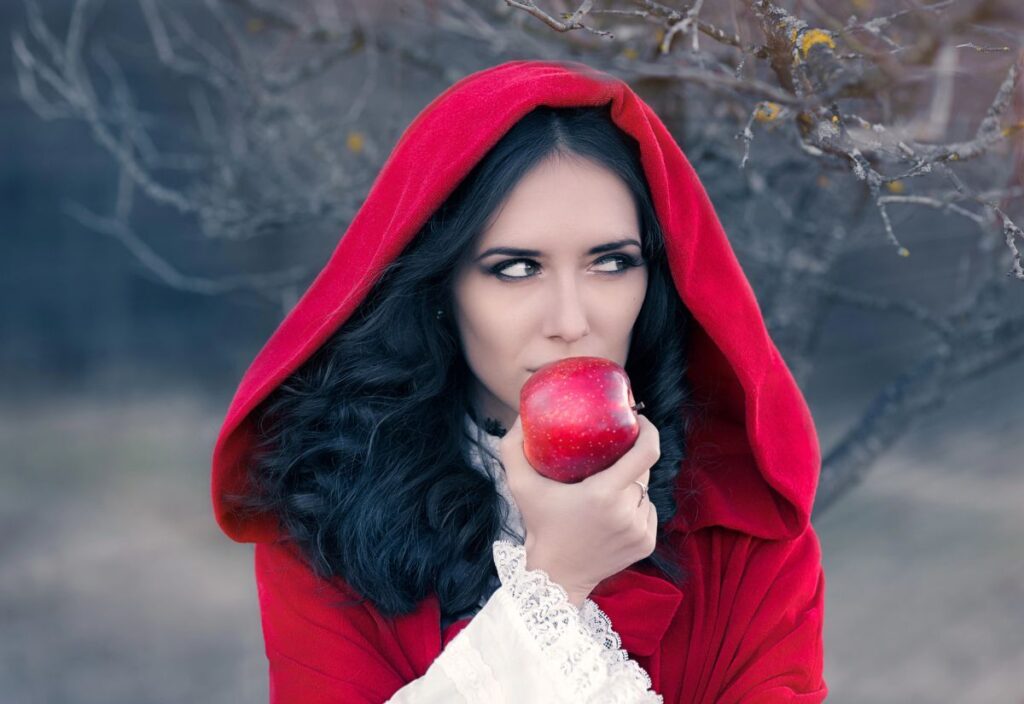 Woman in a red hooded cloak holding a red apple near her mouth, standing outdoors.