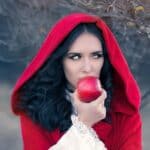 Woman in a red hooded cloak holding a red apple near her mouth, standing outdoors.