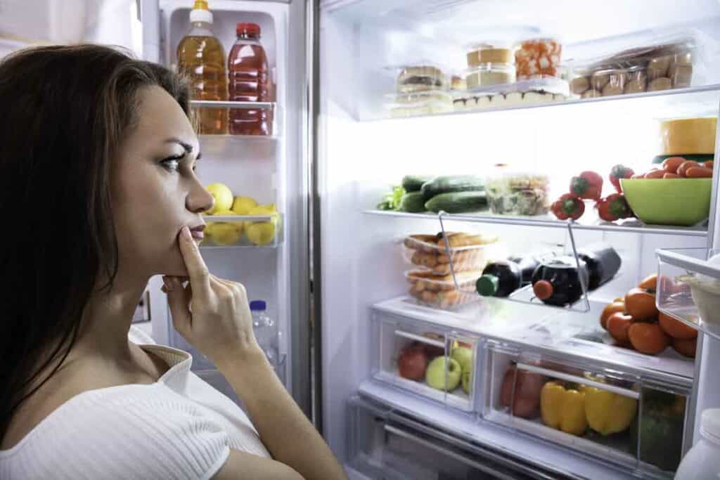 A woman stands in front of an open refrigerator, contemplating what to choose from various fruits, vegetables, and drinks inside.