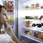 A woman stands in front of an open refrigerator, contemplating what to choose from various fruits, vegetables, and drinks inside.