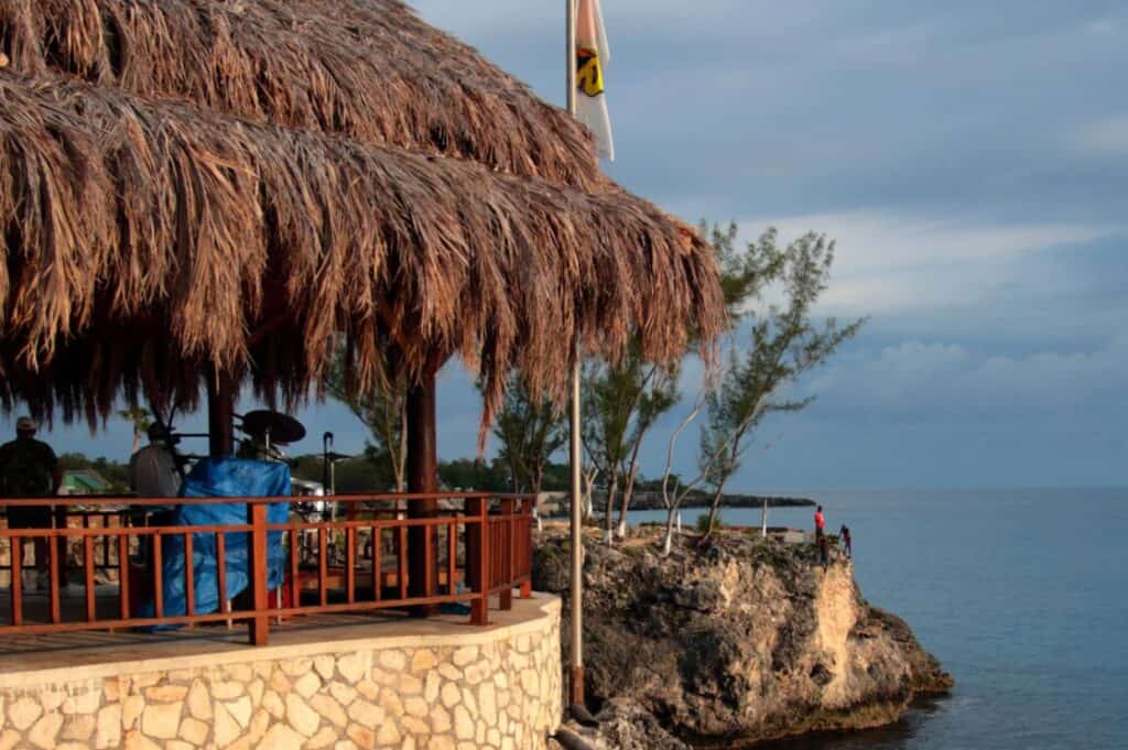 A thatched-roof pavilion, reminiscent of the best beach bars in the Caribbean, overlooks a rocky cliff and the ocean. Two people stand near the edge of the cliff, with trees visible in the background under a cloudy sky.