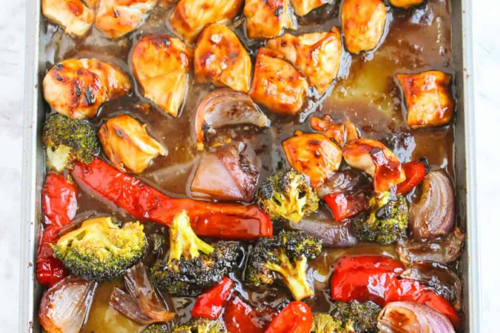 Tray of roasted chicken pieces, red peppers, broccoli, and red onions coated in a glossy sauce.