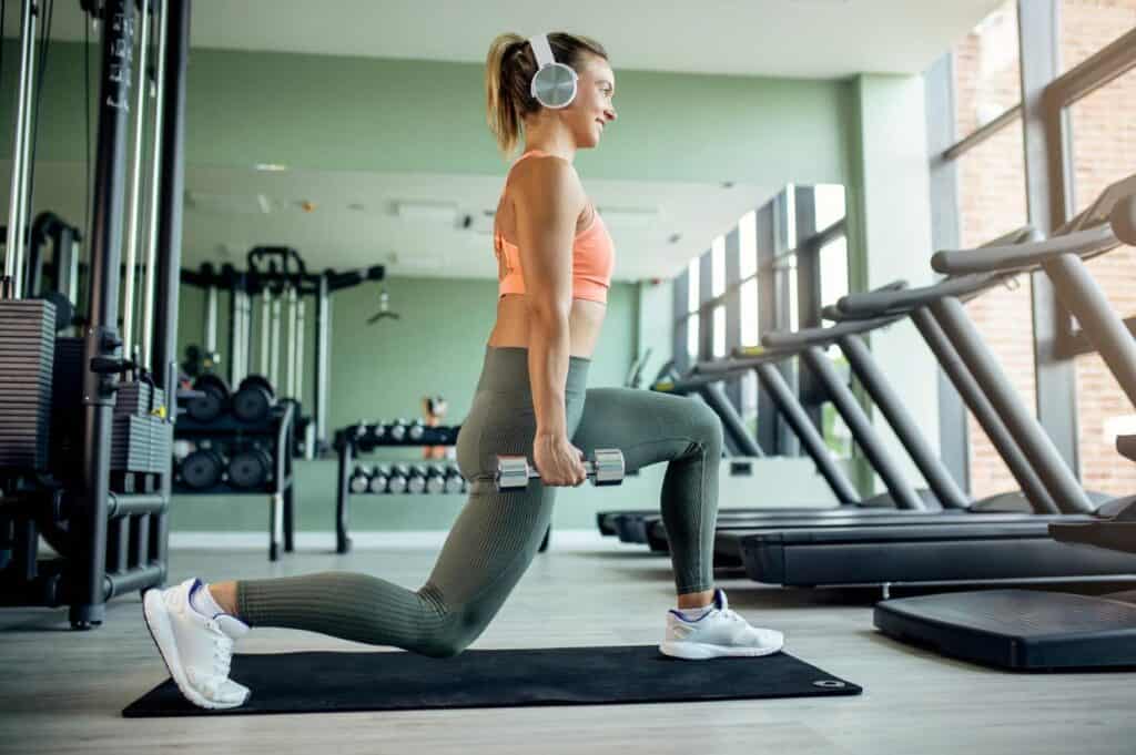 A woman in athletic wear engages in a 20-minute dumbbell workout, performing lunges while wearing headphones and standing on a mat. Fitness equipment is visible in the background.