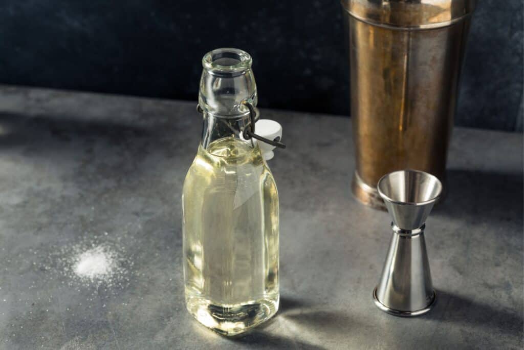 A glass bottle with a metal clasp sits on a countertop next to a metal jigger and cocktail shaker, with a small pile of salt to the side.