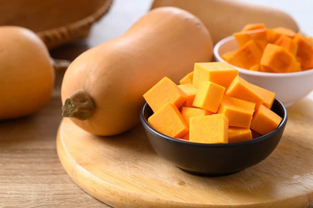 Cubed and whole butternut squash on a wooden board, with bowls containing the cubed pieces.