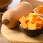 Cubed and whole butternut squash on a wooden board, with bowls containing the cubed pieces.