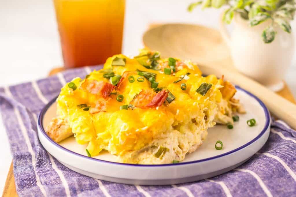 A serving of egg casserole with cheese, bacon, and green onions on a white plate with a blue rim. A glass of iced tea and a wooden spoon are in the background.