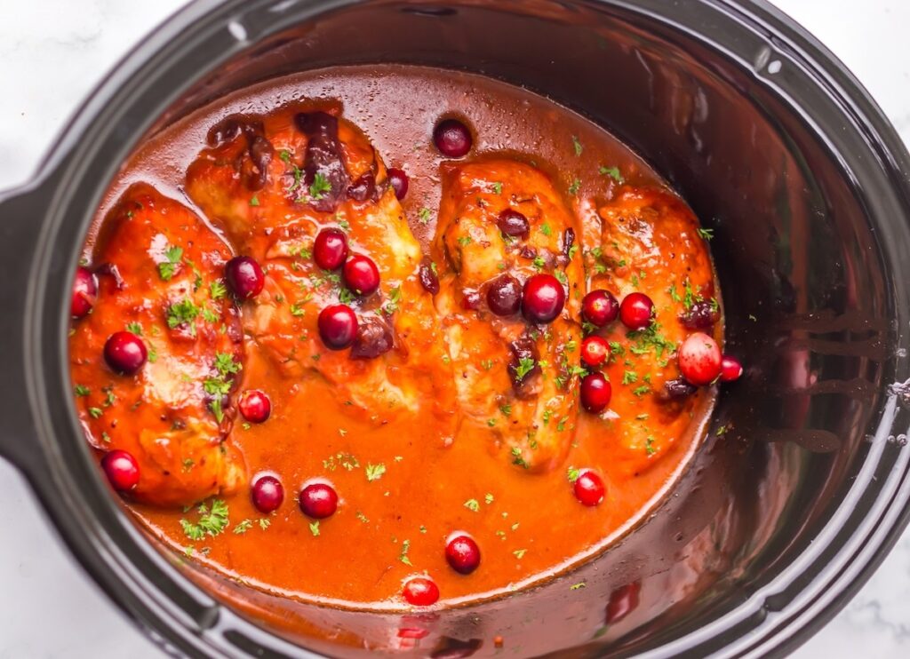 Chicken breasts in a slow cooker with red sauce, cranberries, and herbs.