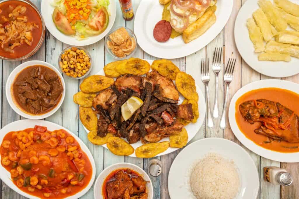 A variety of cooked dishes including meats, plantains, rice, salad, yuca, and sauces, showcasing the culinary delights of the Dominican Republic, arranged on a wooden table with utensils.