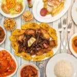 A variety of cooked dishes including meats, plantains, rice, salad, yuca, and sauces, showcasing the culinary delights of the Dominican Republic, arranged on a wooden table with utensils.