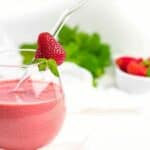 A glass of strawberry smoothie with a straw, garnished with a strawberry and mint. A bowl of strawberries and green leaves are in the background.