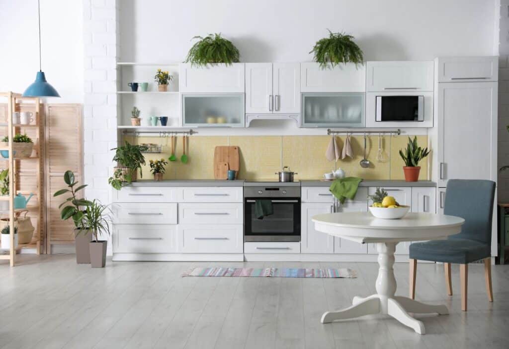 A modern kitchen with white cabinets, green potted plants, a table with a bowl of lemons, and a chair.