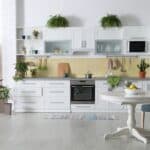 A modern kitchen with white cabinets, green potted plants, a table with a bowl of lemons, and a chair.