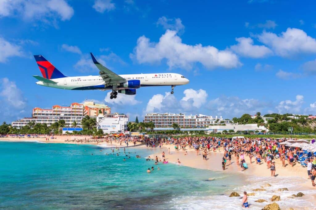 An airplane soars low over a crowded beach near the shore, casting shadows on sunbathers savoring island vibes. In the background, buildings stand tall against a clear blue sky, reminiscent of afternoons spent at the best beach bars in the Caribbean.