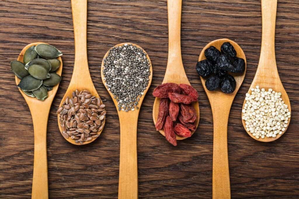 Five wooden spoons on a wooden surface, each holding different seeds and dried fruits: pumpkin seeds, chia seeds, flaxseeds, dried goji berries, dried blueberries, and quinoa.